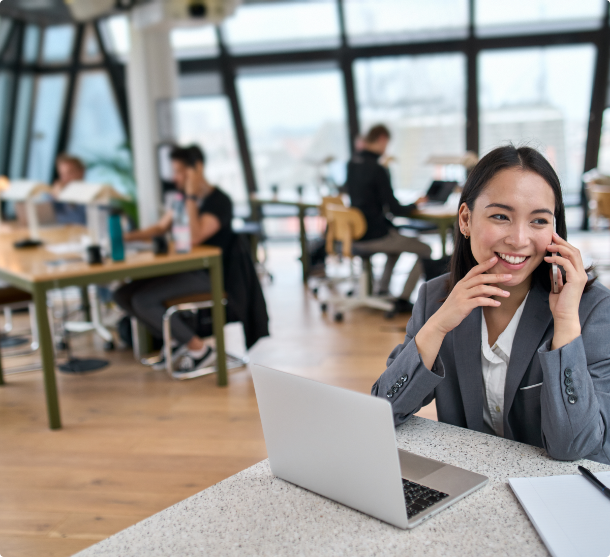 Image of a lady on a phone call sitting at a desk in an open plan office with a laptop, pen and paper in front of her