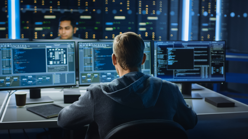 Image of a man sitting at a desk looking at three monitors with code on the screen