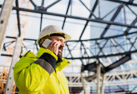 Photo of a man talking on a mobile phone in a construction site wearing a hard hat and a high visibility jacket