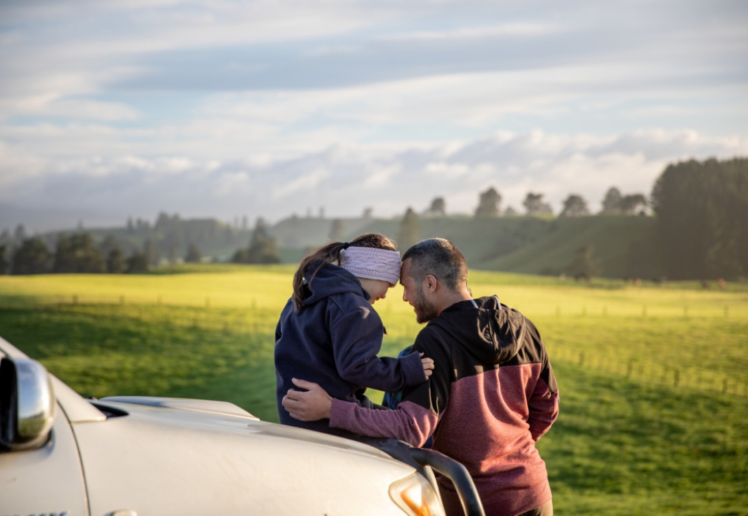 Image of a man and his daughter sitting on the hood of a car looking at each other in front of a green field and mountain range