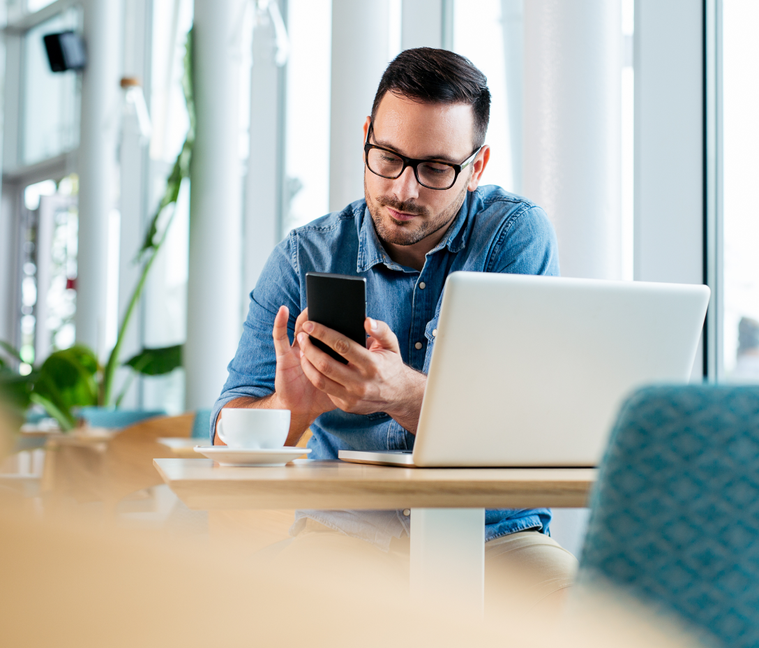 Image of a man sitting at a table with a cup of coffee on his mobile phone and laptop