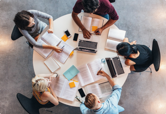 Image of students sitting around a round table with laptops, books, phones an documents studying