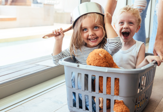 Photo of two children smiling playing in a washing basket with the young girl holding a pot on her head