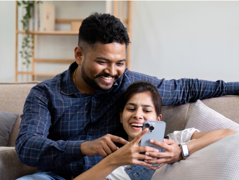 Photo of a couple sitting on the couch with a woman on her phone and her partner looking at the screen over her as they smile at what they are looking at